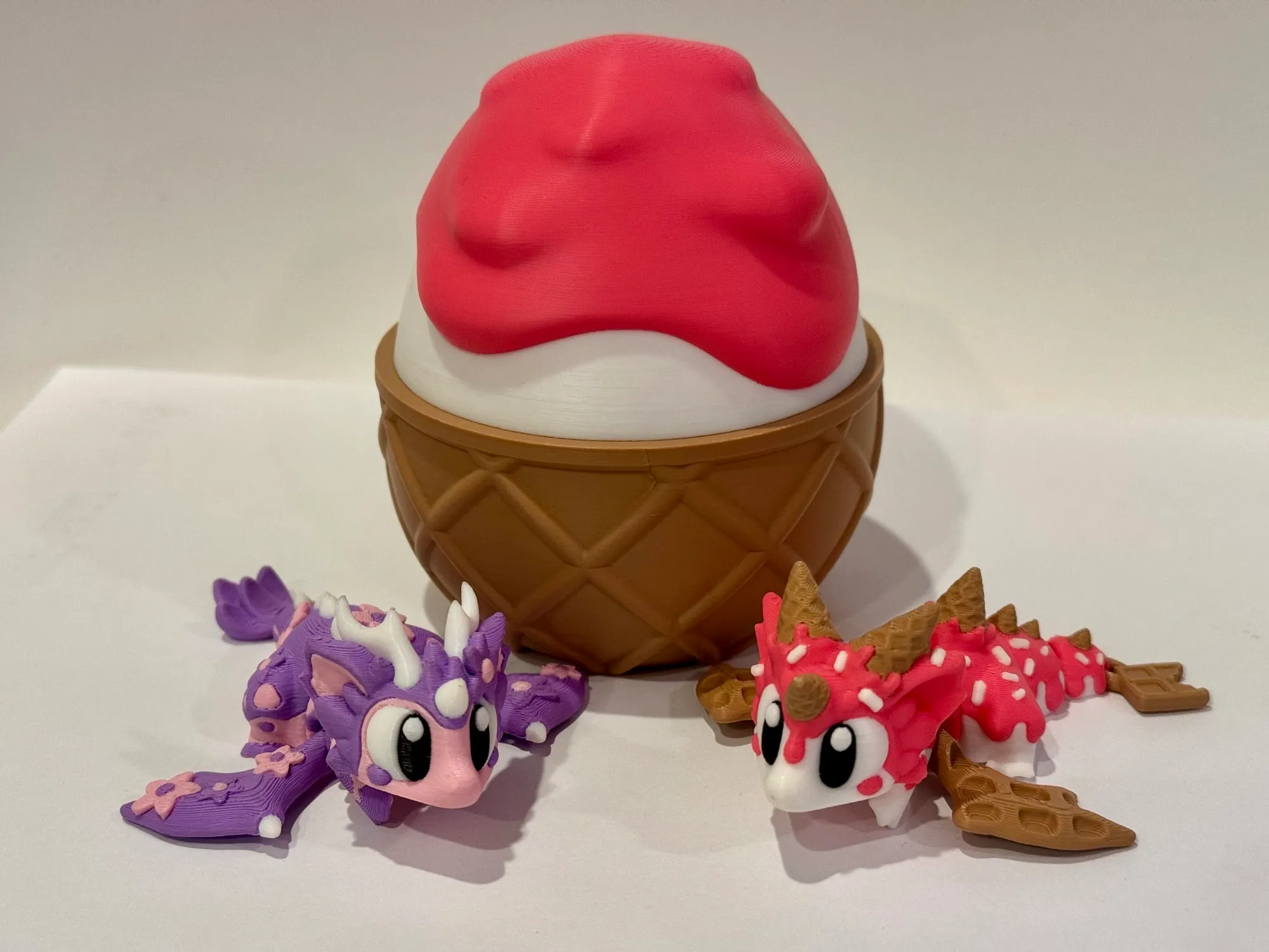 Two articulated 3D printed dragons — one purple with flowers, one strawberry-pink with sprinkles and waffle-cone wings — in front of a large printed ice cream cone.
