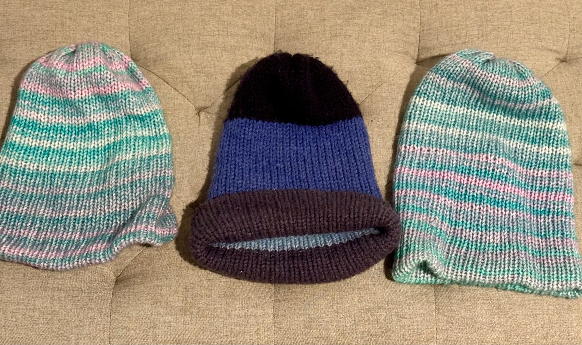 Three hand-knit beanies laid out side by side: a mint-and-pink striped hat, a navy and black roll-brim hat, and a teal and pink striped hat.