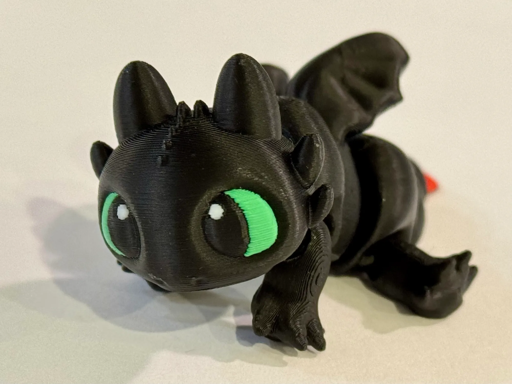 A black 3D printed night-fury dragon with bright green eyes, crouched and looking at the camera.